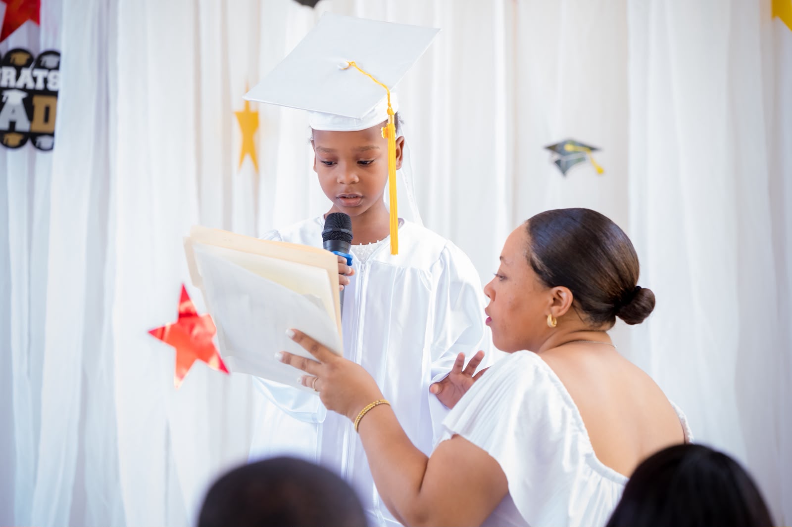 EXL preschool graduate in white cap and gown reading a speech at the microphone with teacher assisting